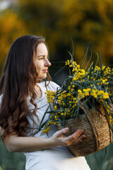 Portrait girl with long hair with a yellow acacia mimosa flower basket. Walk in the flower garden. Girl and flowers. Floristics.Sunset light
