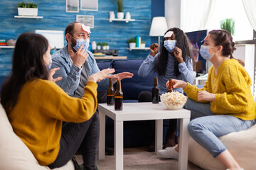 Thoughtful multiethnic friends with sticky notes on foreheads playing name game wearing face mask keeping social distancing preventing covid19 spread drinking beer enjoying popcorn. Conceptual image.
