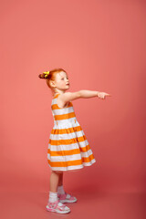 A little girl with red hair points her finger to the left and right. Pink background.