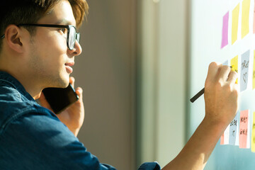 asian adult nature young smart glasses casual cloth work from home hand hold smartphone disscusion remote meeting with another hand write down short note on sticky paper note on window glass