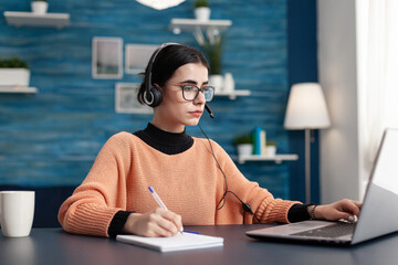 Student with headphones writing notes on notebook while searching communication information using...