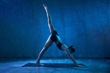 Sporty attractive woman wearing black sportswear standing in yoga asana with raised leg under rain....