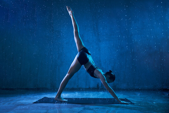 Sporty Attractive Woman Wearing Black Sportswear Standing In Yoga Asana With Raised Leg Under Rain. Strong Flexible Girl Standing In Downward Facing Dog, Adho Mukha Svanasana Pose.