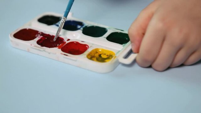 Child painting Closeup. The palette of colors is on the blue table. A child's hand holds a brush and mokes it in red High quality FullHD footage