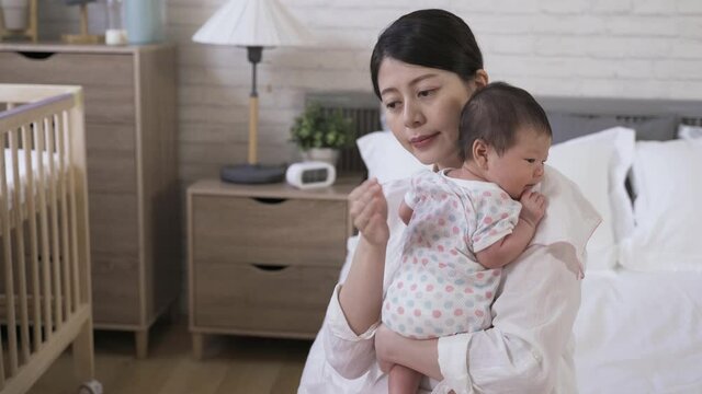 portrait asian mother is helping her adorable newborn to burp with gentle pat on the back in the bedroom at home.