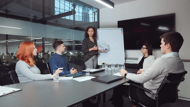Black woman reporting on progress while standing by flipchart with hand-drawn growth graph at business meeting. Multiethnic team analyzing financial success and planning further strategy
