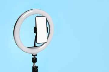 Modern tripod with ring light and smartphone on light blue background. Space for text