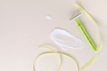 Top view of smear of hair removal cream, green razor and decorative ribbon on the ight beige surface.Empty space 