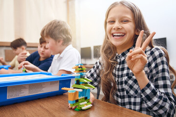 Selective focus of emotional smiling caucasian girl showing piece, working on project, using colorful parts of building kit for toys, creating robot on table. Concept of science engineering.