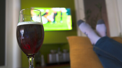 A man in relax watching a soccer match. A glass of dark beer in the foreground. Legs, white socks...