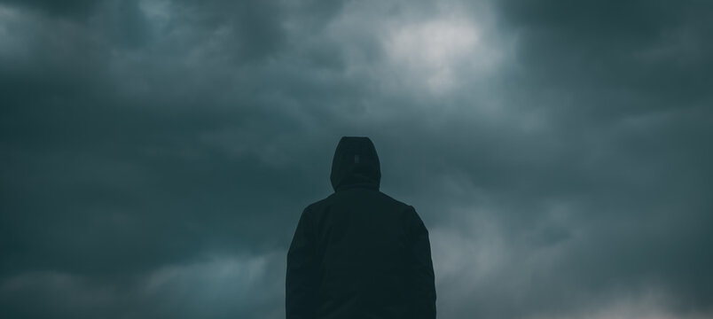 Rear view of male person wearing hooded jacket against dark moody dramatic clouds at sky