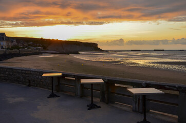Fototapeta na wymiar The beach of Arromanches-les-Bains in Normandy on the coast of the English Channel at sunset, empty tables off the stone wall on the boardwalk, one of the allied landing places on D-Day (Gold Beach)
