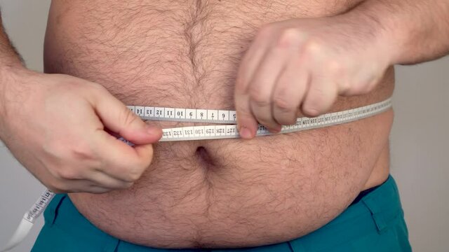 fat guy measures the size of the abdomen with a centimeter measuring tape. the person suffers from excess weight. man with a large and hairy belly