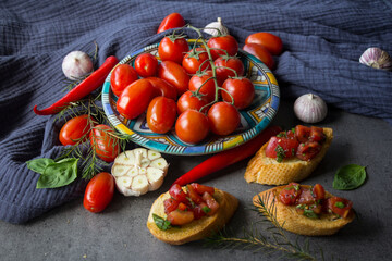 Traditional Italian bruschetta, cherry tomatoes on blue plate, garlic bulbs, basil, pepper and herbs on a table. Top view photo of fresh ingredients on gray textured background. 