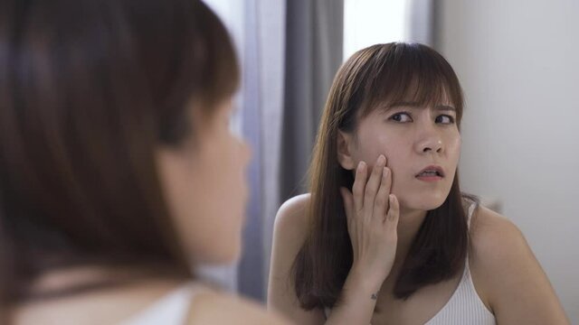 view from behind narcissistic Asian lady looking at her own reflection in the mirror and shocked to find out a zit on her face