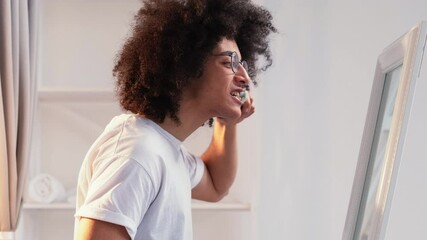 Dental hygiene. Oral health. Morning routine. Profile portrait of confident handsome guy with dark long curly hair brushing teeth with toothbrush looking in mirror in light bathroom.