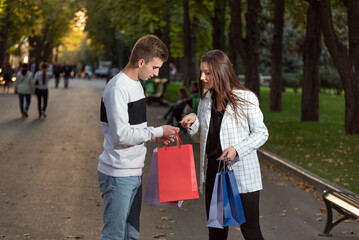 Girl looks into the guys purchases while walking in park. Young couple with shopping bags