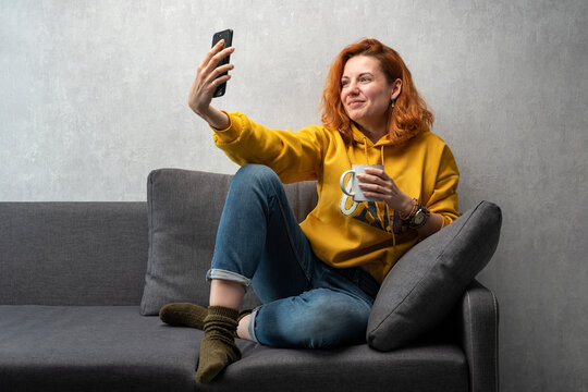 Cheerful young girl sits on the sofa and films herself on camera. Chatting video call. Home selfie