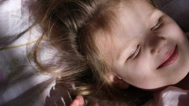 Little girl wake up  lying on comfortable bed with soft pillows at home in dark bedroom with light from window. enjoy happy fun in morning on holiday