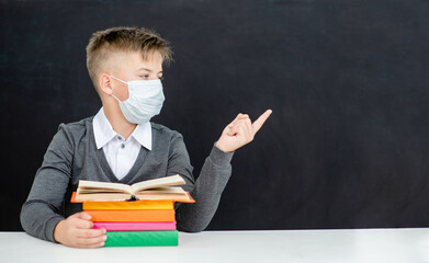 Teen boy wearing protective face mask sits with books at school near blackboatd and points away on empty space