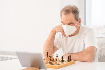 Pensive senior man wearing protective face mask uses tablet computer to play chess with his friends during quarantine Coronavirus (Covid-19) epidemic