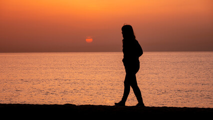 Silhouette of a woman walking by the ocean. Woman shilouette at sunrise on the beach