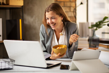 Businesswoman in office having healthy snack. Young woman eating fruit while having a video call.