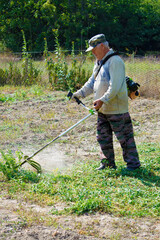 An old man mows grass with gasoline trimmer. Sunny autumn day. Front view.