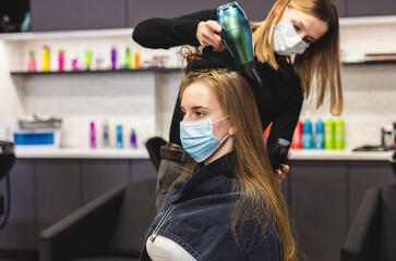 Master woman hairdresser in a medical mask dries the girl's hair with a hairdryer and combs after washing in a beauty salon. Covid-19 pandemic and remedies