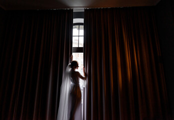 Side view of elegant bride in a peignoir opens the curtains in a hotel room. Concept of mornings bride
