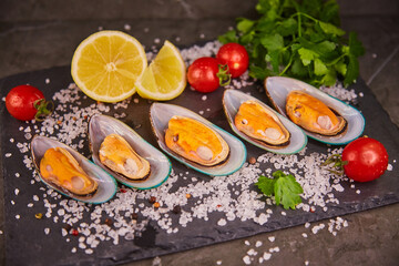 Fresh mussels in a shell lie with parsley and lemon and cherry tomatoes, on a black slate board, on a dark gray table, sea salt is poured around the shells. 