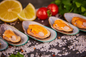 Obraz na płótnie Canvas Fresh mussels in a shell lie with parsley and lemon and cherry tomatoes, on a black slate board, on a dark gray table, sea salt is poured around the shells. 