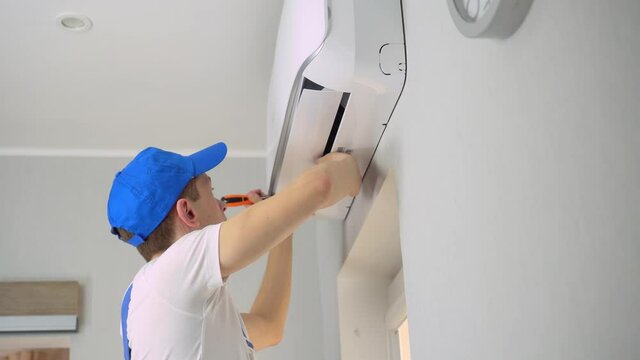 A young master in uniform, blue overalls and a cap, a white T-shirt, installs or repairs the air conditioner in the apartment room.