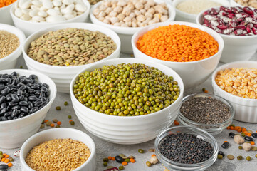 Mix of legumes, chickpeas, lentils, beans, peas, quinoa, sesame, chia, flax seeds in bowls on a gray concrete background.