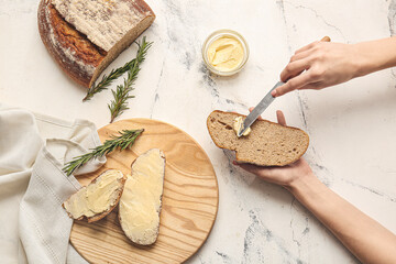 Woman spreading butter onto slice of fresh bread on light background