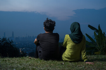 Portrait of Young Asian couple, man and woman, sitting on grass enjoying Kuala Lumpur view during blue hour.