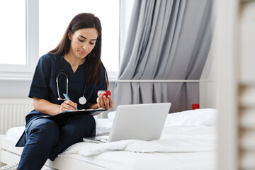 The woman doctor sitting on the bed in front of a laptop writing something on paper while holding a jar of pills in her hand