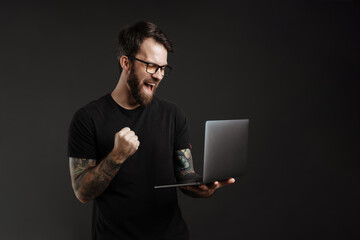 Bearded excited man in eyeglasses making winner gesture and using laptop