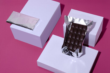Black chocolate bars wrapped in a foil, white boxes on the pink surface
