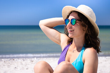 Beautiful brunette girl smiling relaxing on summer beach, sunbathing wearing blue bikini, sunglasses, straw hat. Enjoying sea vacation, life in paradise. Advertising for travel company and agency