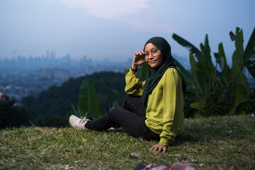 Pretty girl sitting on grass, relaxing, enjoying Kuala Lumpur city view during sunset. Portrait of young Asian woman, wearing hijab and green attire.