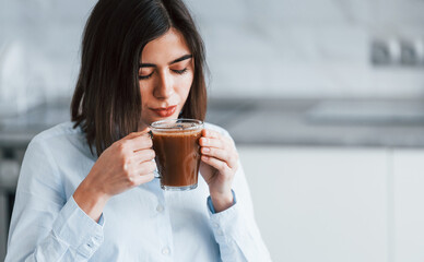 With cup of coffee. Young woman is indoors in room of modern house at daytime