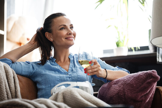Leisure time concept. Happy beautiful woman drinks white wine from glass sitting on a couch indoors. Female spending her free day and relaxing at home alone.