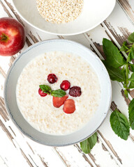 Oatmeal with milk and berries. Nutritious breakfast, wholesome healthy food. Bowl of porridge cereal oats with fresh red berries and mint on on white wooden rustic table. High angle view.
