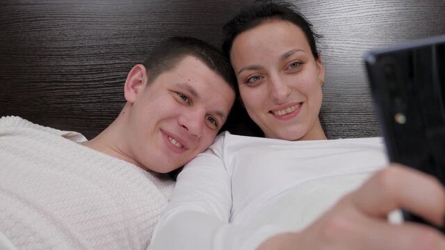 A young couple holding a smartphone in hand is photographed together, shoots video blog on bed. Happy family husband and wife take photos online using a modern mobile phone at home on sofa in bedroom.