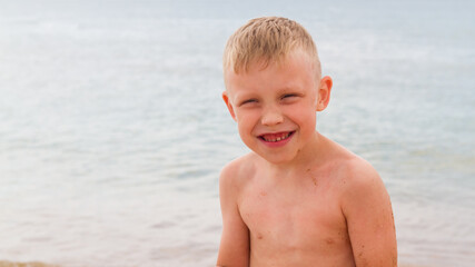 Funny caucasian boy sits on the beach by the sea in summer and laughs