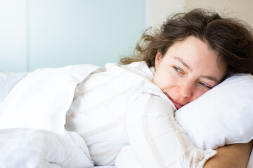 Happy brunette woman lying in bed hugging soft white pillow. Smiling looking in distance, enjoying fresh soft bedding and orthopedic mattress in bedroom. Relaxing, resting, lazy weekend morning.