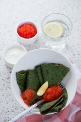 White bowl with spinach crepes, red caviar and sour cream, studio shot on a beige stone background, selective focus
