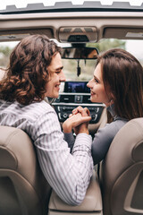 Happy young couple hand in hand in front of car, modern car interior, looking at each other and smiling