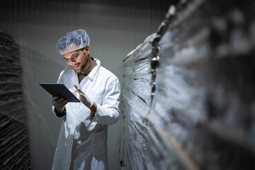Factory worker with hairnet and hygienic gloves holding tablet computer and checking inventory in food cold storage.